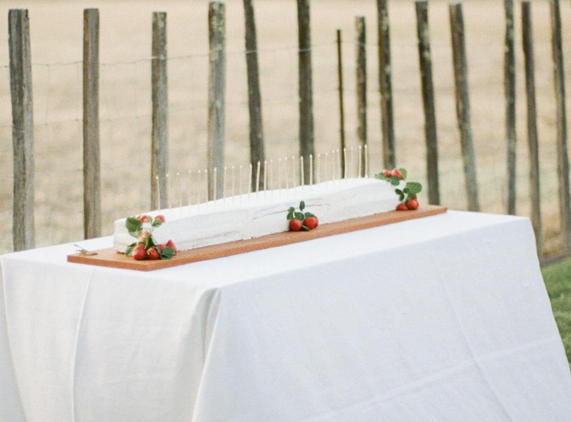 A long celebration cake on a wooden board set on a white tablecloth outdoors, with strawberries and candles